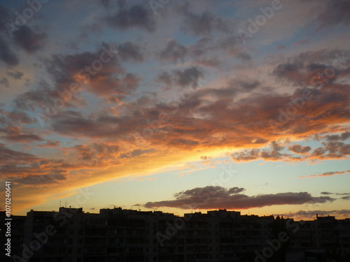 Bright unusual sunset city landscape orange pink rays above cityscape. Brutalist panel buildings appear as black silhouettes against a fiery orange-yellow sky dramatic lighting dark rain storm clouds