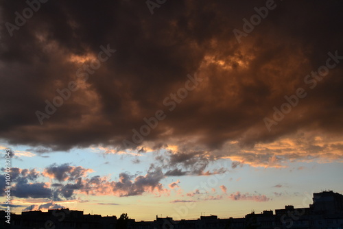 Bright unusual sunset city landscape orange pink rays above cityscape. Brutalist panel buildings appear as black silhouettes against a fiery orange-yellow sky dramatic lighting dark rain storm clouds