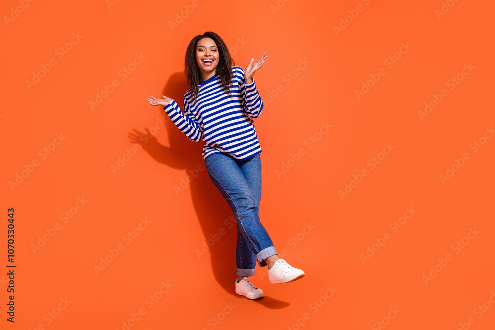 Full length photo of cheerful impressed lady dressed striped shirt walking rising arms hands empty space isolated orange color background Full length photo of cheerful impressed lady dressed striped shirt walking rising arms hands empty space isolated orange color background