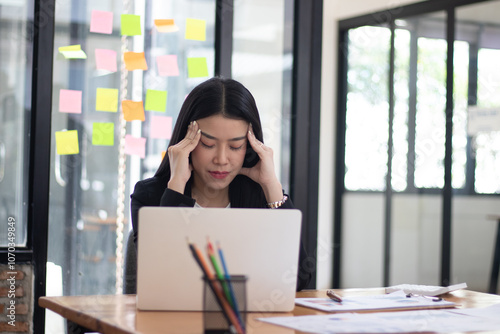 Asian female entrepreneur with headache sitting at company desk