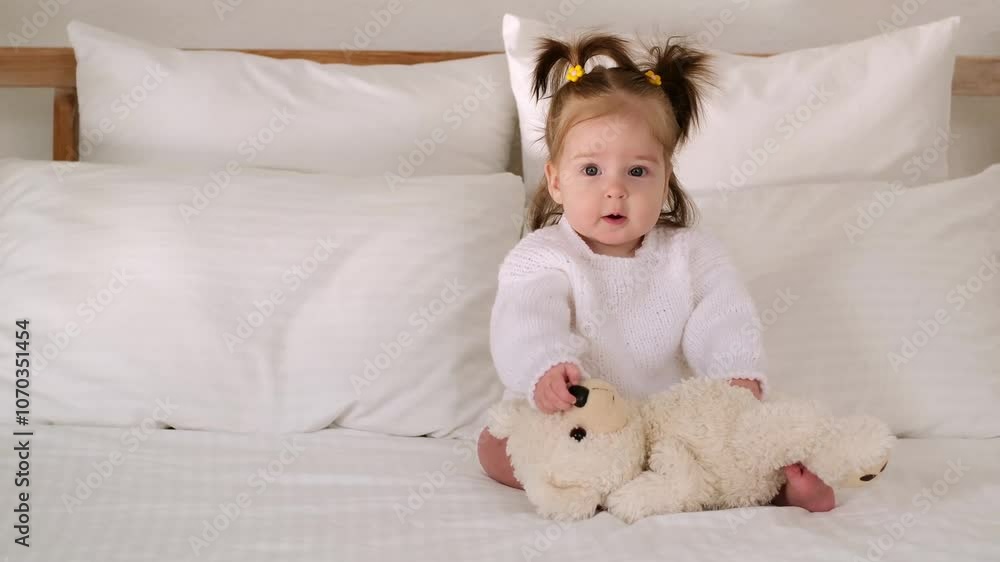 Baby girl on the bed in the bedroom with a stuffed toy smiling or laughing, cute little baby at home in a white sweatshirt Baby girl on the bed in the bedroom with a stuffed toy smiling or laughing, cute little baby at home in a white sweatshirt