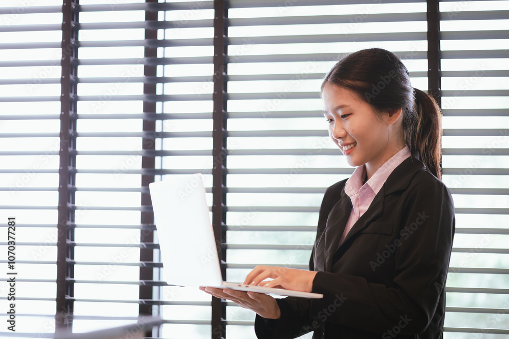 Young Asian woman in suit using laptop by window in office. Close-up of body. Copy space available. Young Asian woman in suit using laptop by window in office. Close-up of body. Copy space available.