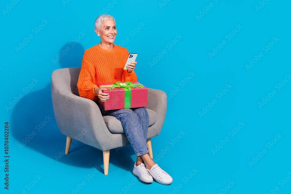 Photo of old lady sit chair use cell smart device look empty space deliver gift box for christmas isolated blue color background Photo of old lady sit chair use cell smart device look empty space deliver gift box for christmas isolated blue color background