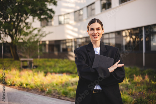 Woman entrepreneur with ponytail crossed hands hold tablet is standing on office building background. Successful smiling caucasian woman in business suit. Female worker with crossed arms look happy.