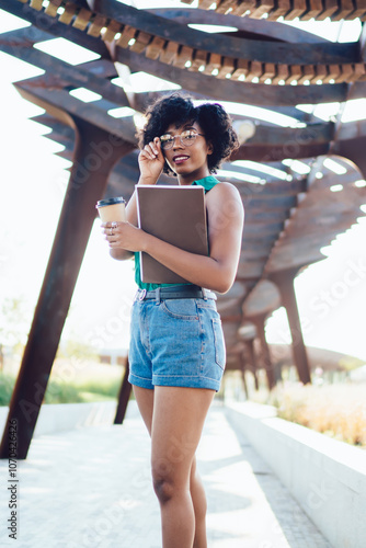 Full length portrait of trendy dressed hipster girl with curly hair standing on street, dark skinned pretty young woman in spectacles looking at camera  spending time on urban setting architecture #1070426426