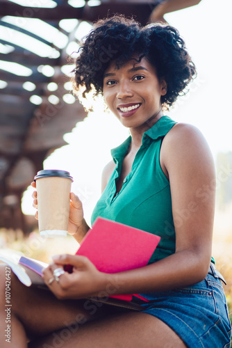 Portrait of african american woman with curly hair sitting with book in city park recreating on summer weekends, beautiful dark skinned hipster girl holding coffee to go cup looking at camera #1070426427