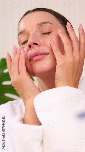 happy woman applying serum or cream on face, during facial ritual at home.smiling girl with natural skin using massage roller.double side mirror and cosmetic jars in front,monstera leaf as decor.