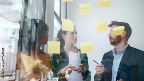 Video of successful business team writing on stickers at office glass board while discussing together in the coworking space.