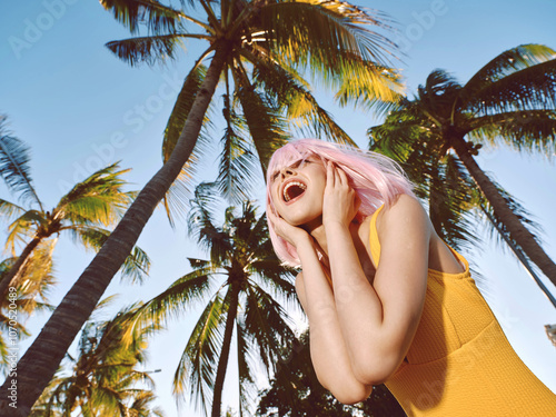 Pinkhaired woman smiling, standing in front of palm trees with hands on head, tropical background, joyful expression, casual summer outfit, outdoor portrait, relaxed vacation mood photo