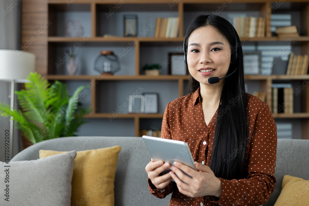 Asian woman with headset using tablet at home, smiling, seated on couch. Concept of remote work, online communication, technology. Ideal for virtual meeting, customer service, or e-learning. Asian woman with headset using tablet at home, smiling, seated on couch. Concept of remote work, online communication, technology. Ideal for virtual meeting, customer service, or e-learning.