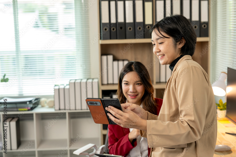 Obraz premium Two businesswomen sharing ideas and smiling while looking at smartphone in office