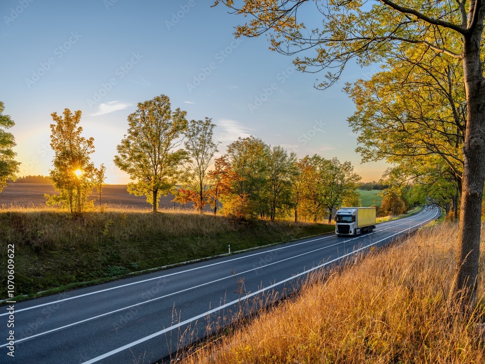 Silver truck driving on an asphalt road between rows of leafy trees in autumn colors in a rural landscape at sunset