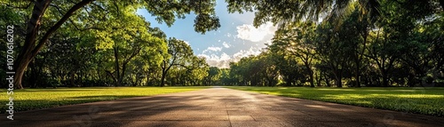 Sunlit park with empty paved area, framed by lush trees and green lawns, blue sky with clouds, serene atmosphere, urban natural escape