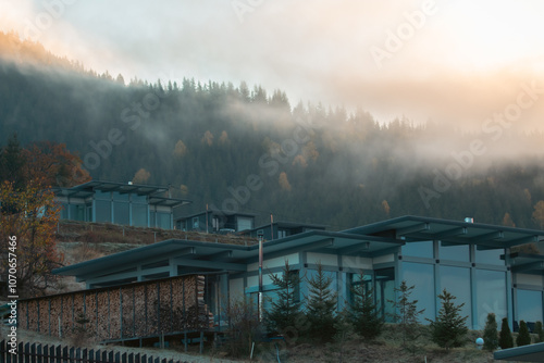 Luxury houses in autumn mountains covered with clouds in the morning. Modern hotel in Carpathian mountains, Ukraine. Foggy morning in mountain resort. House in autumn forest on foggy hills. 