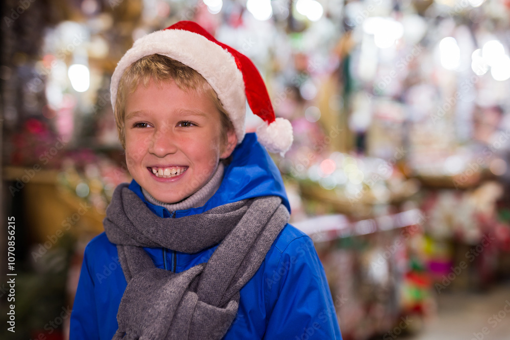 Cheerful laughing tween boy wearing warm scarf and Santa hat standing outdoors on blurred background of colorful Christmas market.... Cheerful laughing tween boy wearing warm scarf and Santa hat standing outdoors on blurred background of colorful Christmas market....