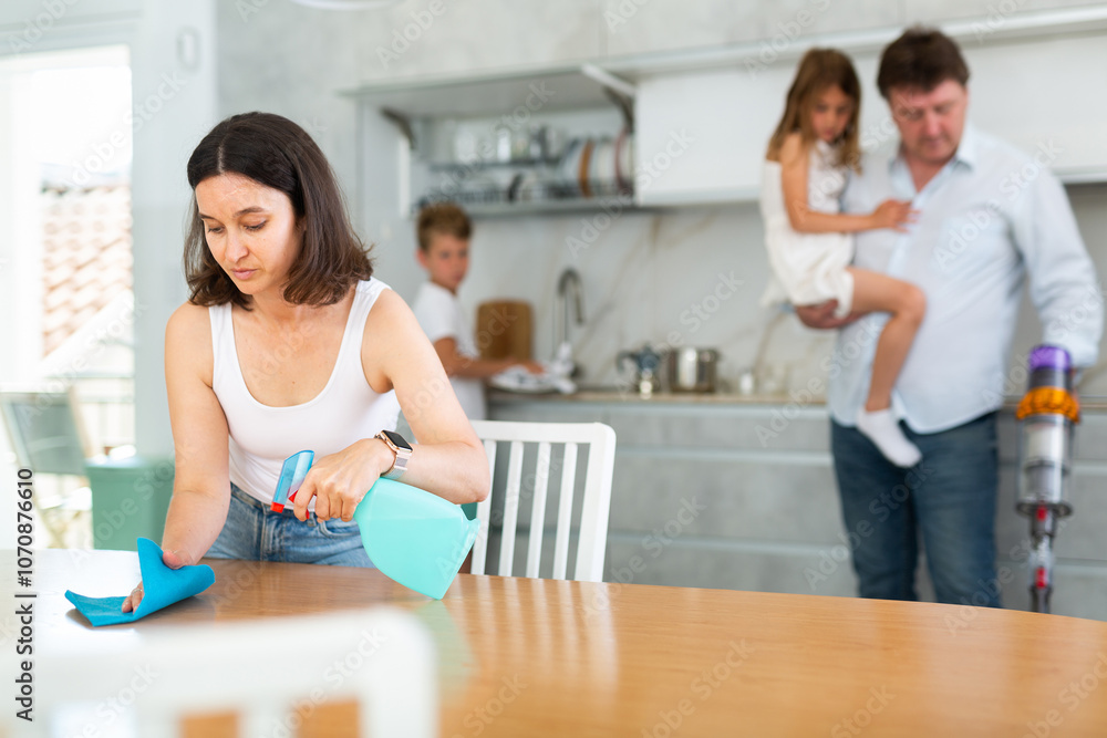 Joint family house cleaning, household tasks, household help. Mom wipes kitchen table, husband holds little daughter in arms and vacuums floor in background, blurred. Joint family house cleaning, household tasks, household help. Mom wipes kitchen table, husband holds little daughter in arms and vacuums floor in background, blurred.