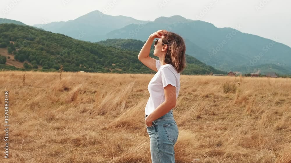 A woman joyfully stands in a vast field, appreciating the serene beauty of nature and majestic mountains A woman joyfully stands in a vast field, appreciating the serene beauty of nature and majestic mountains