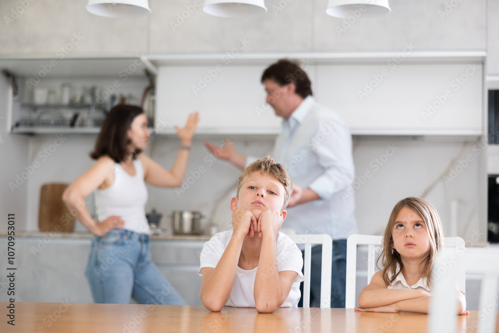 Two children sitting at the table sadly and their mother and father bickering behind in the kitchen Two children sitting at the table sadly and their mother and father bickering behind in the kitchen