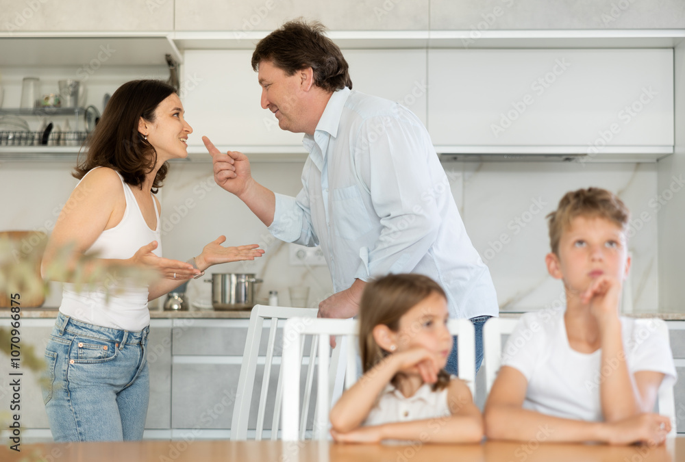 Angry parents quarrelling in the kitchen and bored preteen brother and little sister sitting at the table Angry parents quarrelling in the kitchen and bored preteen brother and little sister sitting at the table