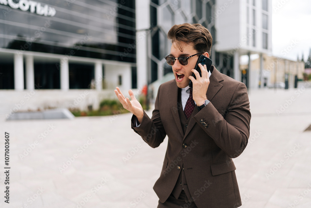 Portrait of mad screaming businessman in sunglasses and suit yelling on smartphone outside standing by office building, gesturing angrily with one hand. Concept of business communication. Portrait of mad screaming businessman in sunglasses and suit yelling on smartphone outside standing by office building, gesturing angrily with one hand. Concept of business communication.