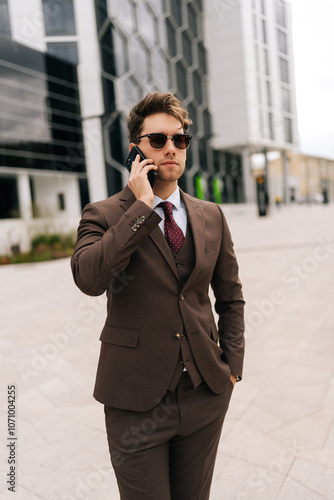 Vertical portrait of successful businessman in suit and sunglasses making call using mobile phone standing in city street on cloudy summer day, exuding style. Concept of business communication