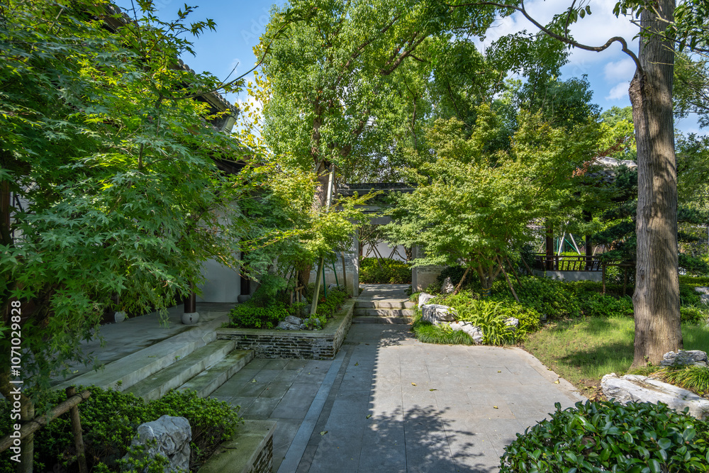 A tranquil stone pathway winds through a lush traditional Chinese garden, featuring mature trees providing dappled shade, manicured shrubs, rocks and landscaping elements. A tranquil stone pathway winds through a lush traditional Chinese garden, featuring mature trees providing dappled shade, manicured shrubs, rocks and landscaping elements.