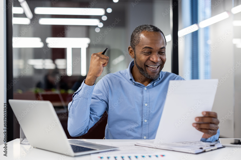 Mature african american businessman seated at office desk, celebrating success. Smiling joyfully while holding documents, pen, and using laptop. Symbolizes professional achievement and happiness. Mature african american businessman seated at office desk, celebrating success. Smiling joyfully while holding documents, pen, and using laptop. Symbolizes professional achievement and happiness.