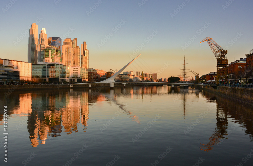 Fototapeta premium Cityscape of Puerto Madero neighborhood with Puente de La Mujer or Woman's Bridge, High-rise buildings, harbour and river during sunset in Buenos Aires, Argentina. With copy space