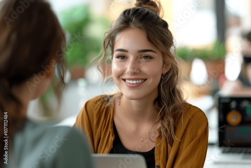 Happy young woman talking with female coworker at desk, smiling and working on laptop, office meeting, good vibes, business education, internship, training, professional skills, mentoring