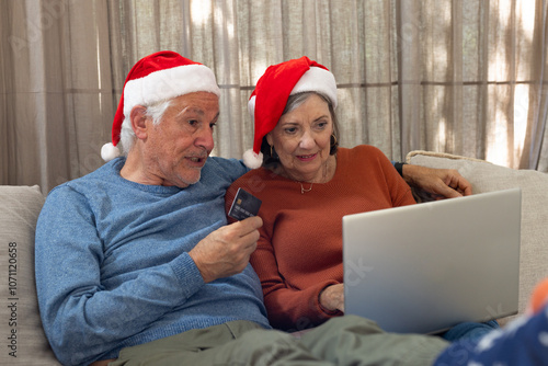 Christmas time, Senior couple wearing Santa hats shopping online with laptop at home photo