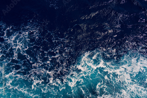 Aerial shot capturing the contrast between dark ocean waves and bright white seafoam, creating dynamic and dramatic seascape. Backgrounds or marine themes