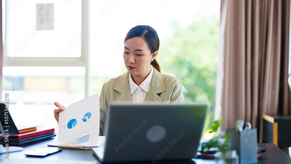 A professional woman in a light suit sits at a desk, focused on her work with documents and a laptop in front of her. The bright office setting A professional woman in a light suit sits at a desk, focused on her work with documents and a laptop in front of her. The bright office setting