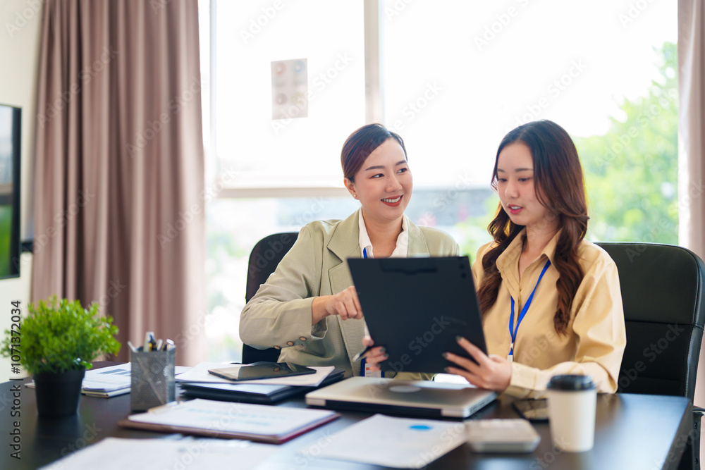 Two businesswomen in formal attire are discussing work in a modern office. One points to the laptop screen while the other observes, focused on a collaborative task Two businesswomen in formal attire are discussing work in a modern office. One points to the laptop screen while the other observes, focused on a collaborative task