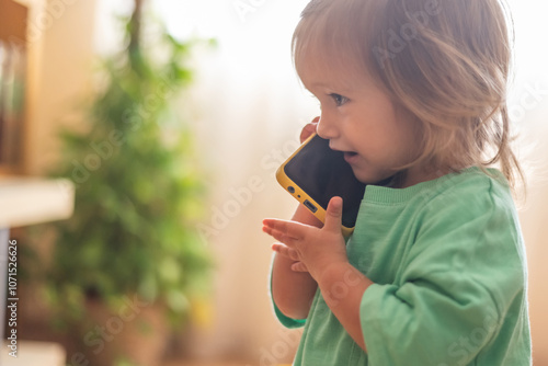 Cute Baby Pretending To Talk On Mobile Phone, Smiling And Imitating Adult Behavior In A Genuine Way.Authentic Expression photo