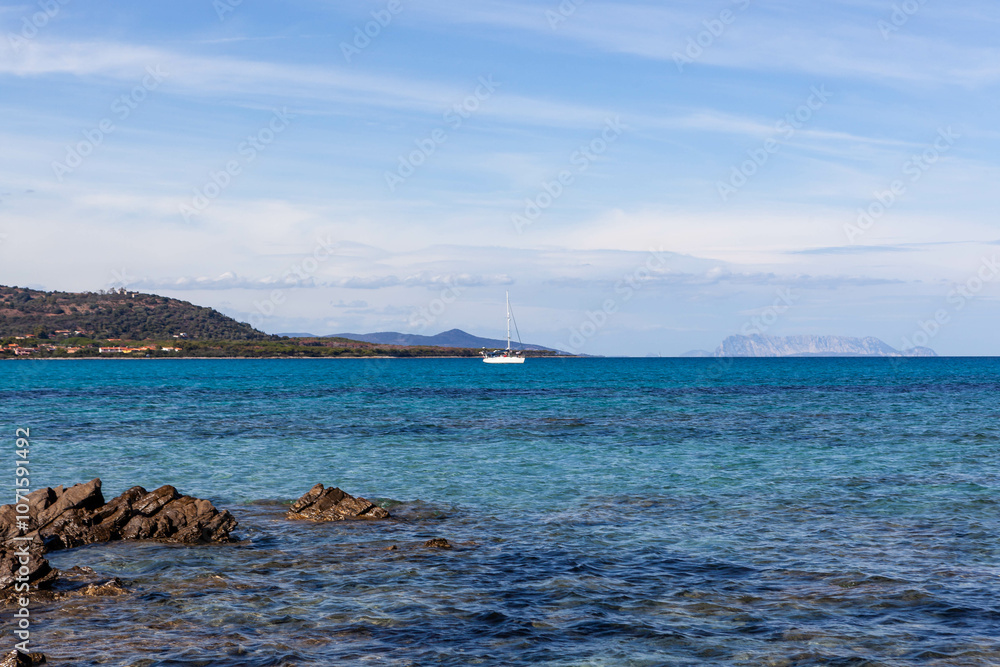 Crystal clear turquoise emerald Mediterranean sea and white sailboat in the middle. Eastern coast of Sardinia and island Tavolara in the background. Crystal clear turquoise emerald Mediterranean sea and white sailboat in the middle. Eastern coast of Sardinia and island Tavolara in the background.