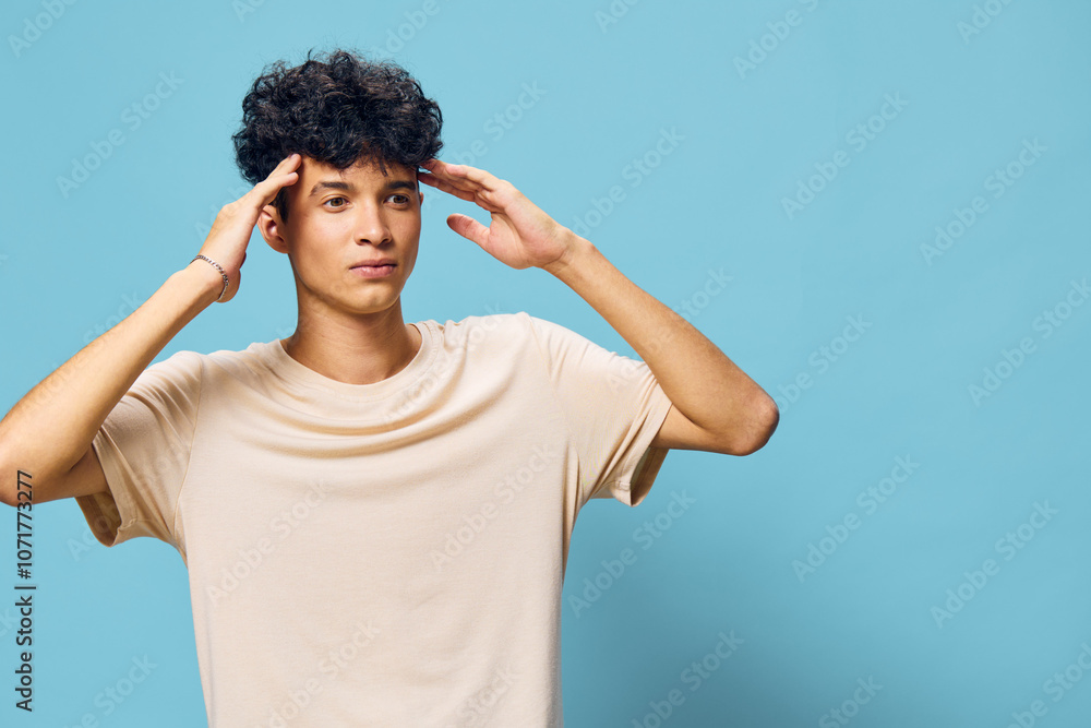 Young man with curly hair holding his head in a thoughtful pose, wearing a beige t shirt against a light blue background, conveying a sense of contemplation or stress Young man with curly hair holding his head in a thoughtful pose, wearing a beige t shirt against a light blue background, conveying a sense of contemplation or stress