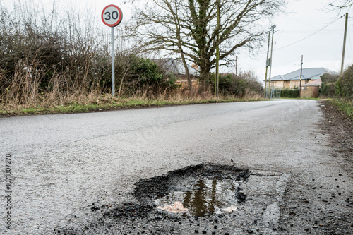 Deep and dangerous pothole seen on a typical British rural road which causes damage to vehicle wheels. A 30 mph road speed sign is seen at the entry to the british village area.