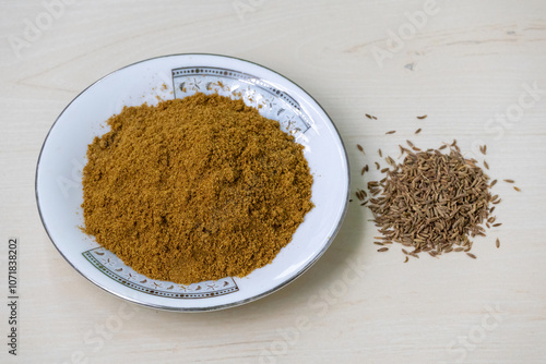 Cumin seed powder is piled on a plate, with a small pile of whole cumin seeds kept beside. Cumin seed powder is a versatile spice with a warm, earthy flavor. It is also known as jeera, jira, or zeera. photo