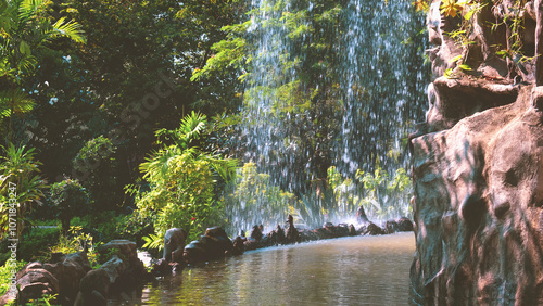 Large artificial waterfall and stone decoration with many greenery plants in public park garden, beautiful outdoor evergreen natural landscape view background