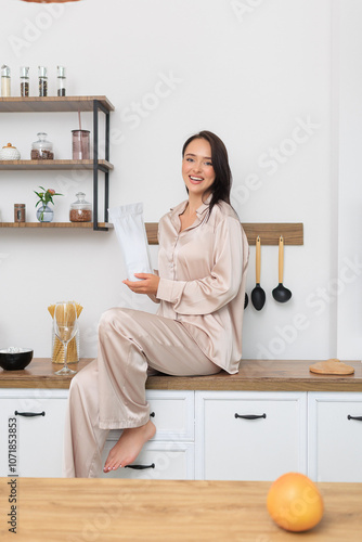 Smiling woman in cozy loungewear sitting on a kitchen counter, holding a container. Modern, bright kitchen setting with organized shelves and fresh ingredients, embodying a relaxed lifestyle.