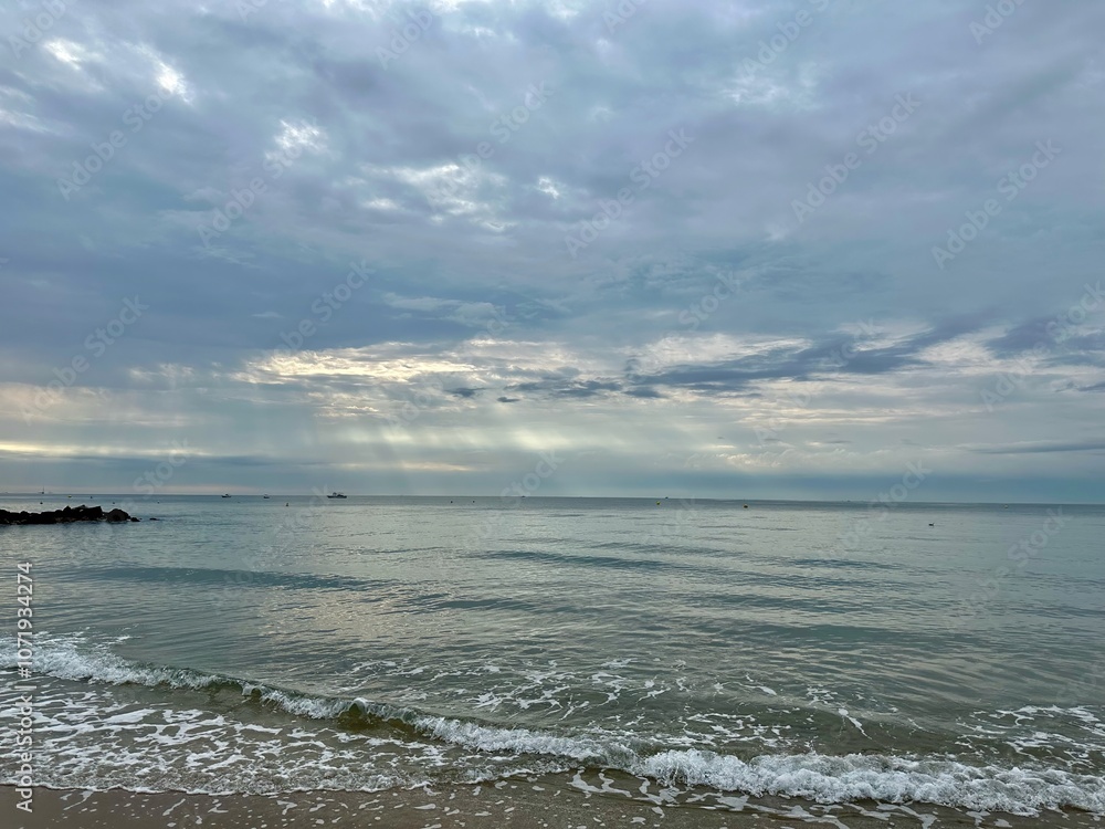 Peaceful seascape at dawn with soft waves and a distant boat under a dramatic, cloudy sky on Palavas-les-Flots beach, Mediterranean Sea, France Peaceful seascape at dawn with soft waves and a distant boat under a dramatic, cloudy sky on Palavas-les-Flots beach, Mediterranean Sea, France