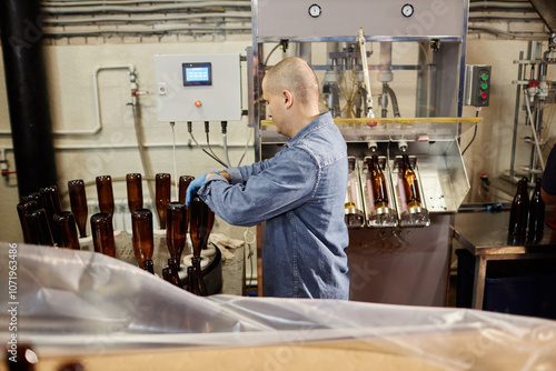 Side view of concentrated male production operator carefully sterilizing glass bottles while directing cleaning machine in workshop of cider or beer factory, copy space photo