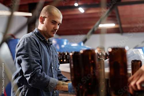 Medium shot of hard working male blue collar worker wearing denim overalls operating production line in workshop of cider or beer factory, copy space photo