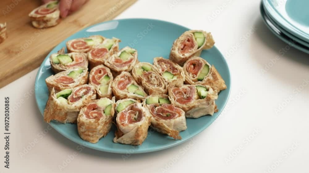 Close-up of hand arranging salmon and cucumber wrap rolls on a blue plate, perfect for appetizers or party snacks Close-up of hand arranging salmon and cucumber wrap rolls on a blue plate, perfect for appetizers or party snacks