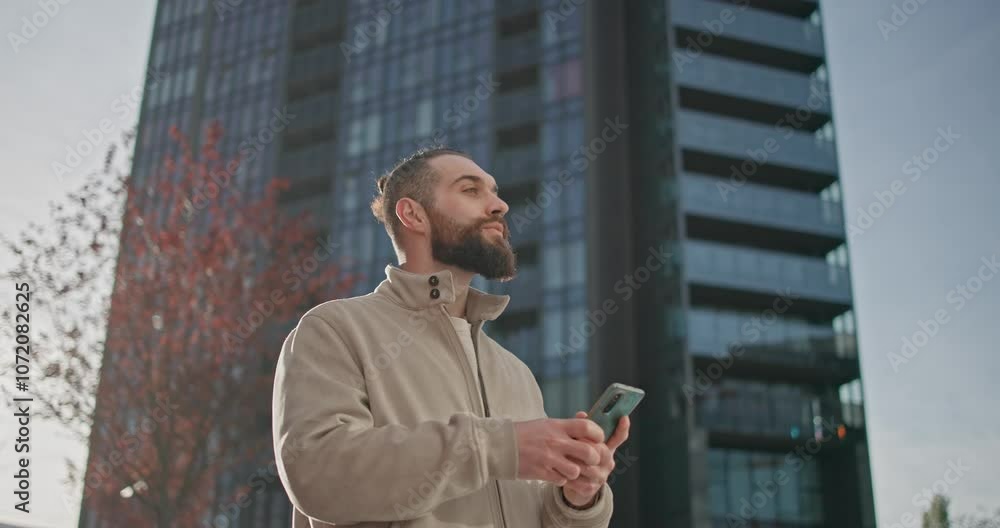 Young man with stylish trendy hairstyle and beard in casual clothes in urban environment standing against modern buildings background and reading messages or news on phone Young man with stylish trendy hairstyle and beard in casual clothes in urban environment standing against modern buildings background and reading messages or news on phone
