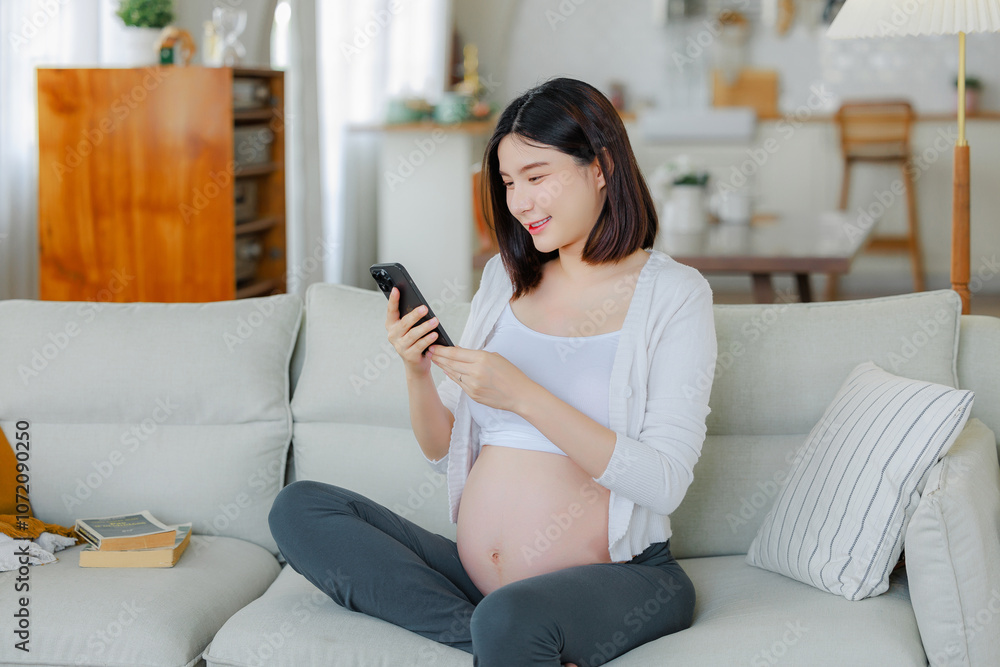 Young Asian happy pregnant woman is sitting and using mobile phone on sofa and touching her belly. Family plan, pregnancy, motherhood, people and expectation concept Young Asian happy pregnant woman is sitting and using mobile phone on sofa and touching her belly. Family plan, pregnancy, motherhood, people and expectation concept