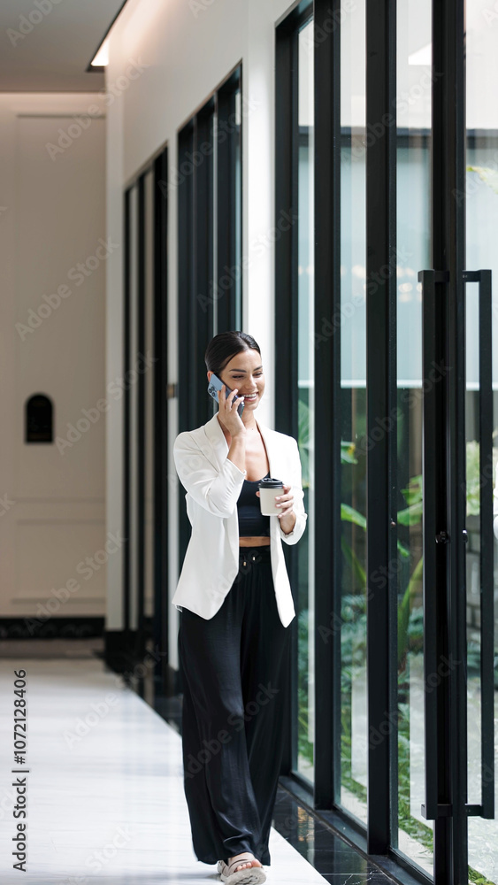 Attractive and confident businesswoman holding a cup of coffee and talking on the phone in the corridor of an office building. Concept of confidence, professionalism and multitasking. Attractive and confident businesswoman holding a cup of coffee and talking on the phone in the corridor of an office building. Concept of confidence, professionalism and multitasking.