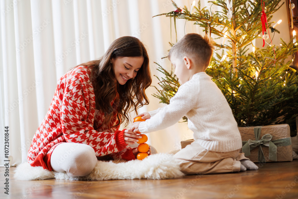 Fototapeta premium Boy playing with tangerines near smiling mother and Christmas tree at home