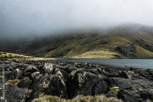 A large body of water sits nestled at the base of a misty, green-covered mountain. The water is calm, reflecting the overcast sky. Scattered rocks line the shoreline.