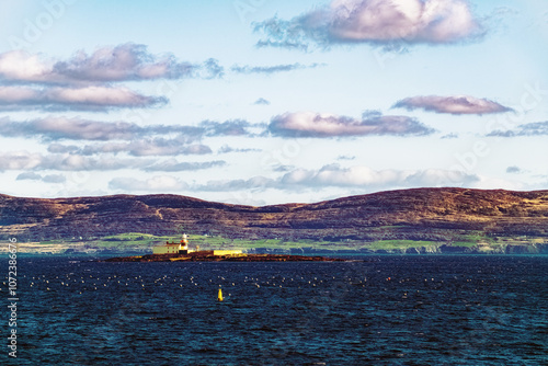 A single lighthouse, standing tall and white against a backdrop of rugged terrain and a vast expanse of ocean water. The structure features a red roof and sits on a small rocky island. photo